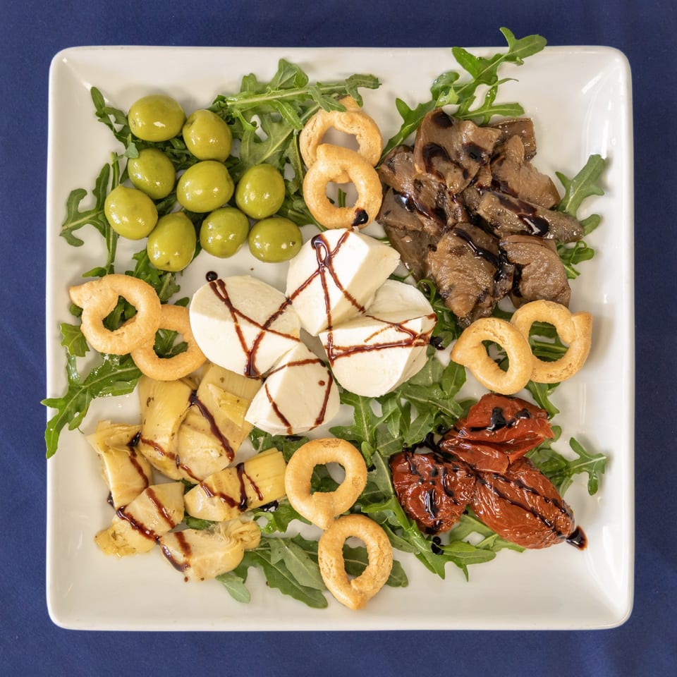 A Milane Italian antipasto platter with olives, mozzarella, artichokes, sun-dried tomatoes, and taralli on arugula.