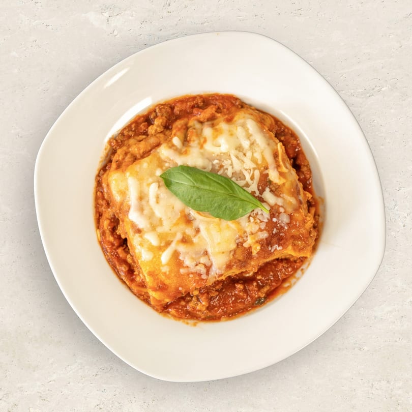A white plate holds a serving of lasagna with melted cheese, red sauce, and a basil leaf.