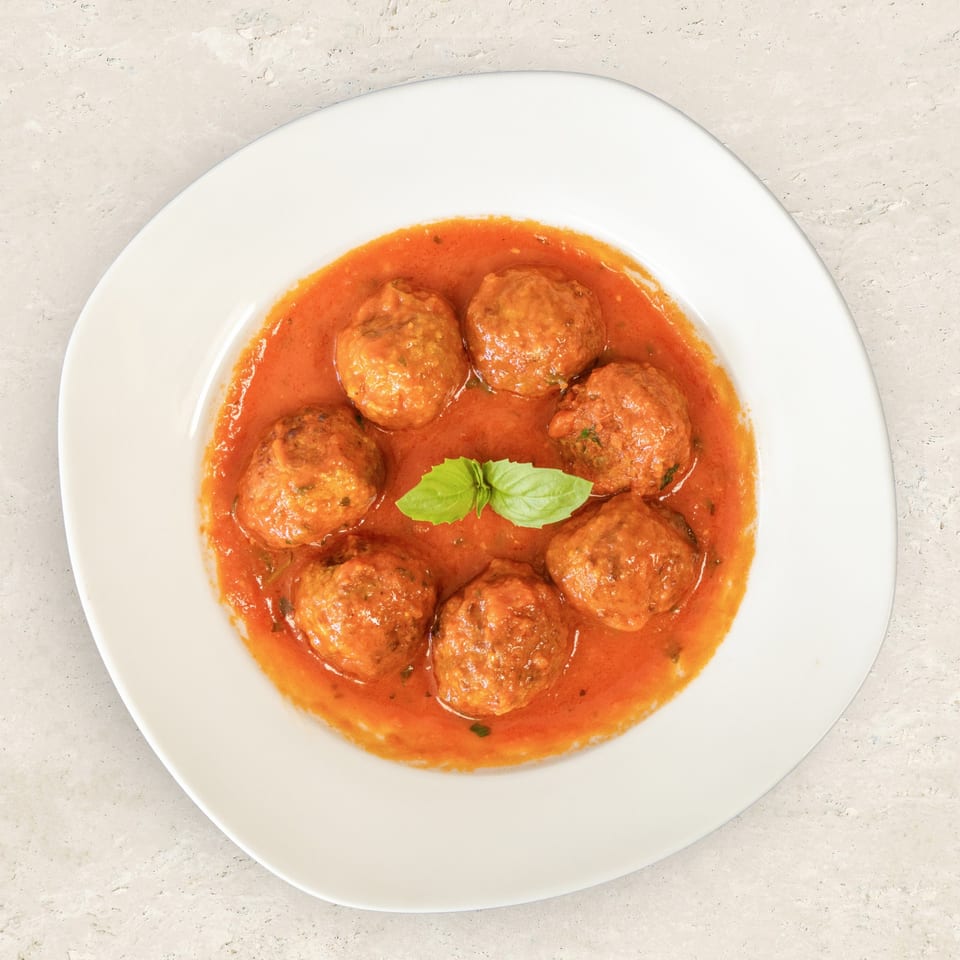 Seven meatballs in a rich red tomato sauce, garnished with fresh basil, on a white plate.